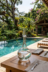 Clear glass decanter on a wooden table by a pool with lush greenery in the background