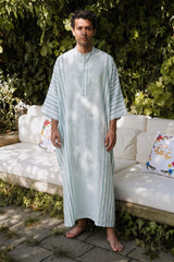 Man wearing a light blue striped jellaba standing outdoors with greenery and a white couch in the background.