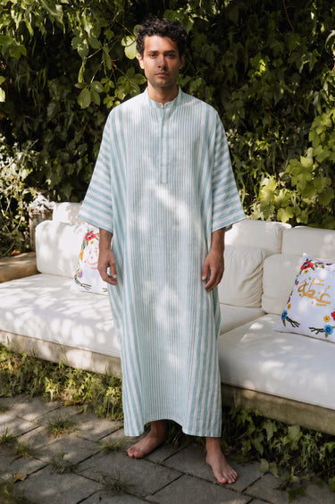 Man wearing a light blue striped jellaba standing outdoors with greenery and a white couch in the background.