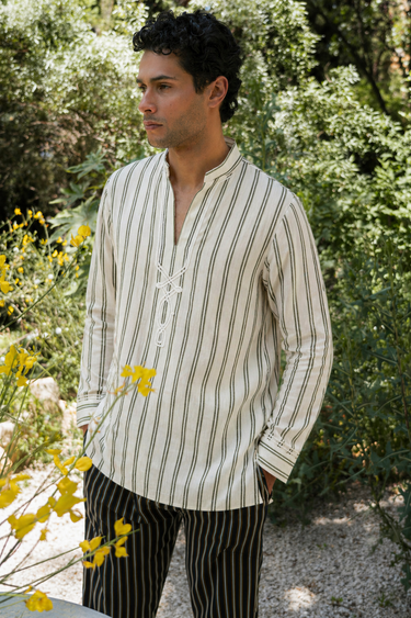 Man wearing a striped shirt and black pants standing outdoors with greenery and flowers in the background