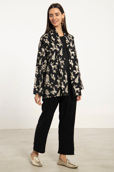 A woman modeling a black long-sleeve blazer with a cream dog pattern, paired with black wide-leg pants and metallic gold loafers, set against a plain white studio background.