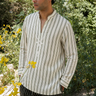 Man wearing a striped shirt and black pants standing outdoors with greenery and flowers in the background