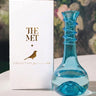 Blue glass bottle with decorative design next to a white box labeled 'The Met' on a blurred floral background