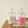 Two glass jars with lids on a wooden table, one containing pink candies and the other white stones.
