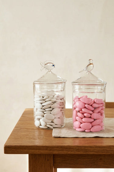 Two glass jars with lids on a wooden table, one containing pink candies and the other white stones.